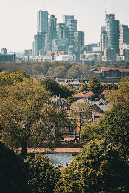 A view of a busy city skyline with numerous modern high-rise buildings and skyscrapers in the background, featuring glass facades reflecting sunlight. In the foreground, there is a park area with lush green trees and a small pond or water feature, where a few people are visible near the water. Inside the park, residential houses with sloped roofs and gardens are seen among the trees. The scene suggests the environment of a residential area transitioning into an urban business district, captured during daytime with clear weather. This image exemplifies the typical setting for house removals and relocation services, highlighting the contrast between domestic homes and commercial high-rises, with Movers Canary Wharf providing professional moving and furniture transport solutions in such environments.