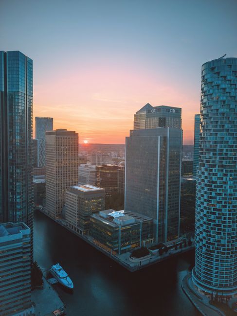 A view of a busy city skyline with numerous modern high-rise buildings and skyscrapers in the background, featuring glass facades reflecting sunlight. In the foreground, there is a park area with lush green trees and a small pond or water feature, where a few people are visible near the water. Inside the park, residential houses with sloped roofs and gardens are seen among the trees. The scene suggests the environment of a residential area transitioning into an urban business district, captured during daytime with clear weather. This image exemplifies the typical setting for house removals and relocation services, highlighting the contrast between domestic homes and commercial high-rises, with Movers Canary Wharf providing professional moving and furniture transport solutions in such environments.