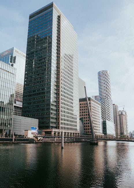 A cluster of modern high-rise office buildings along the waterfront in Canary Wharf with glass façades reflecting the sky, including a prominent tall building with a dark, grid-patterned exterior and a nearby cylindrical skyscraper with a textured white surface. The scene captures the urban environment typical of a major financial district, with the calm water of the river in the foreground and clear, bright weather overhead. This setting is relevant to home relocation and furniture transport services provided by Movers Canary Wharf, often involving loading and unloading furniture or packing materials near these buildings during house removals or moving logistics in the area.
