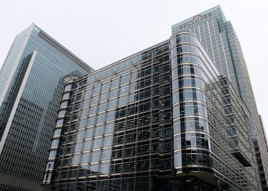 A large modern office building with a curved glass facade, reflecting neighboring skyscrapers in its windows. The building features multiple floors with extensive glass paneling supported by a metal framework, and has a rounded corner on the right side. In the background, there are taller office towers, also made of glass and steel, with one displaying the Citigroup logo near the top. The scene is set outdoors during daylight, with a light overcast sky overhead. This urban environment illustrates the type of commercial property involved in professional house removals and furniture transport services, where careful packing and strategic loading within such high-rise buildings are essential. Movers Canary Wharf utilise appropriate equipment like trolleys and straps to assist with the safe loading process in locations like Heron Quays, ensuring efficient relocation logistics adapted to city centre environments.