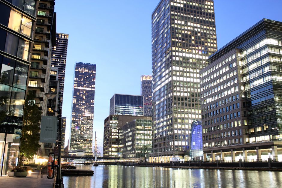 A cluster of modern high-rise office buildings along the waterfront in Canary Wharf with glass façades reflecting the sky, including a prominent tall building with a dark, grid-patterned exterior and a nearby cylindrical skyscraper with a textured white surface. The scene captures the urban environment typical of a major financial district, with the calm water of the river in the foreground and clear, bright weather overhead. This setting is relevant to home relocation and furniture transport services provided by Movers Canary Wharf, often involving loading and unloading furniture or packing materials near these buildings during house removals or moving logistics in the area.