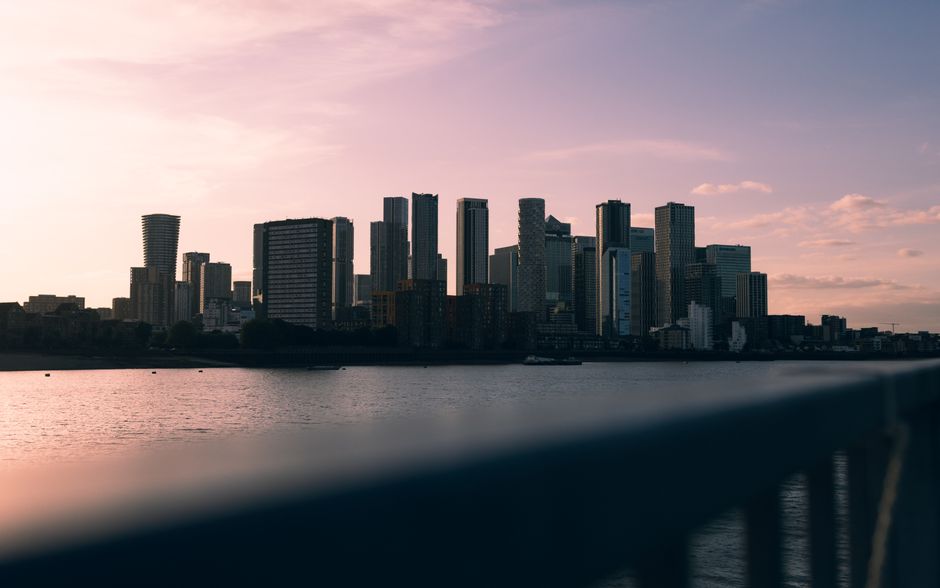 A panoramic view of the Canary Wharf skyline at sunset, featuring numerous modern high-rise buildings, some under construction with cranes visible atop, situated along the riverbank. The buildings are