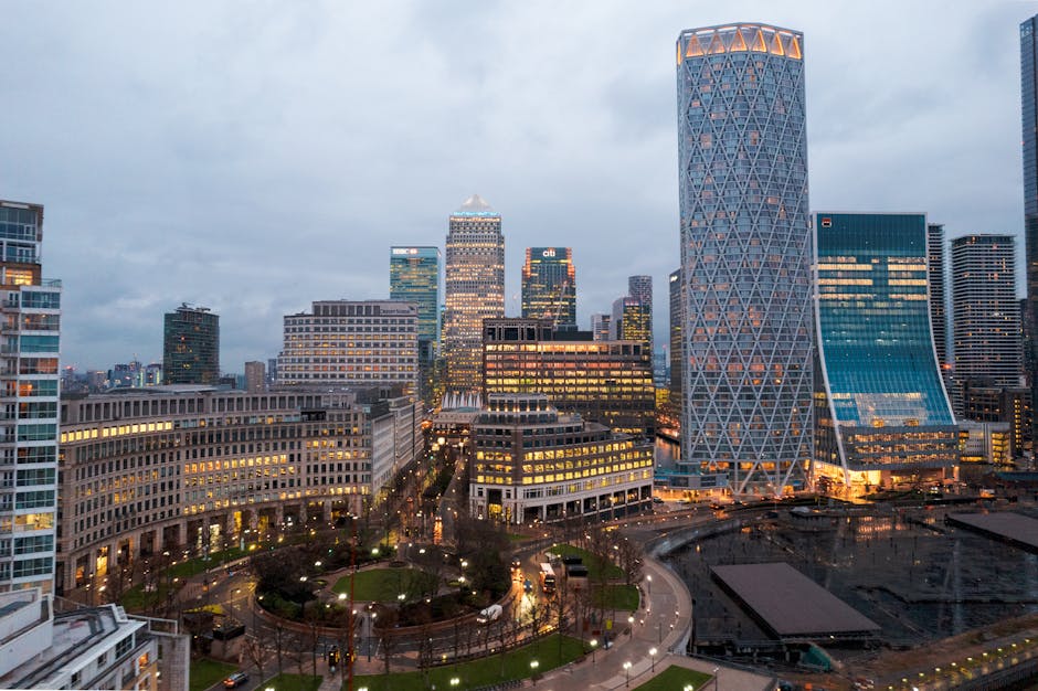 A modern glass office building at Canary Wharf, featuring reflective windows that mirror the partly cloudy sky and surrounding skyscrapers. The building has a curved entrance canopy made of glass and
