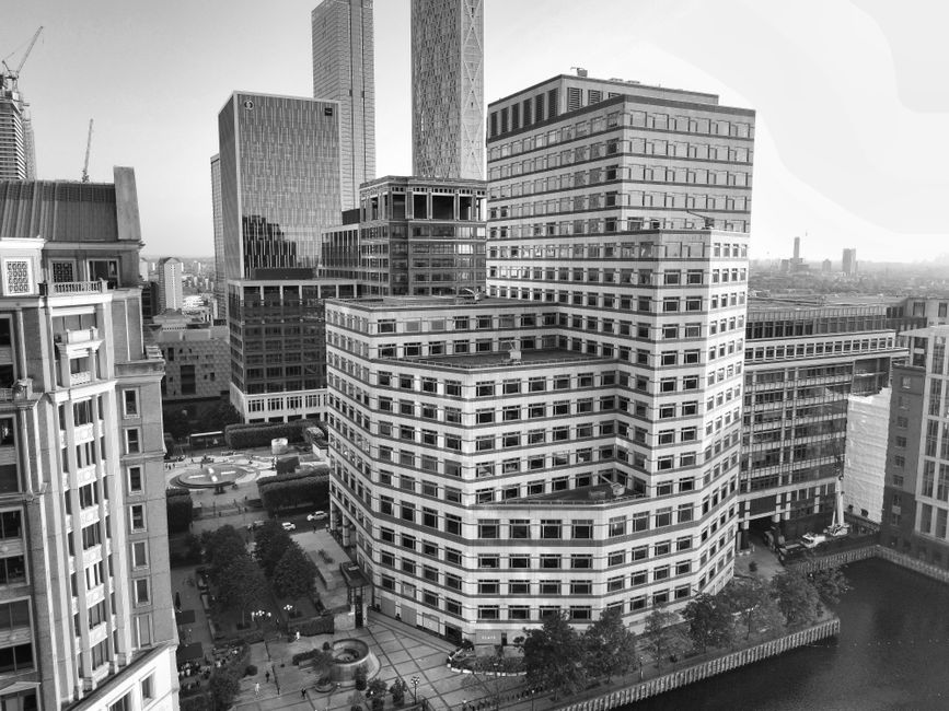 A cityscape scene during dusk featuring tall modern office buildings with illuminated windows reflecting on a waterway at the bottom of the frame. On the left, part of a residential or commercial buil